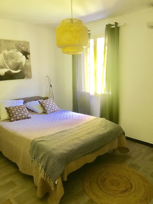 A bedroom with a bed, a hanging light fixture, a window with curtains, and a wall-mounted air conditioning unit.