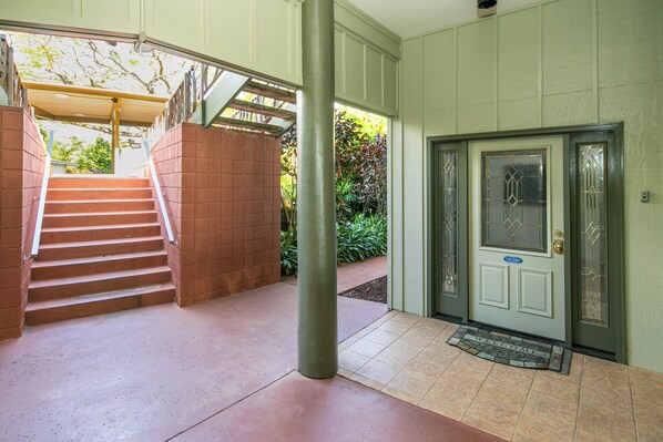 A staircase leading to a door with a doormat, a column, and a view of greenery outside. - Summer Deal Oceanfront Kapaa Condo with Pool and Glass Wall Views (Kapaa)