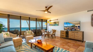 A spacious living room with a large window overlooking the ocean, a ceiling fan, a wooden coffee table, and a television on the wall. - Oceanfront Wailea condo w/ Molokini views and pool access (Wailea)