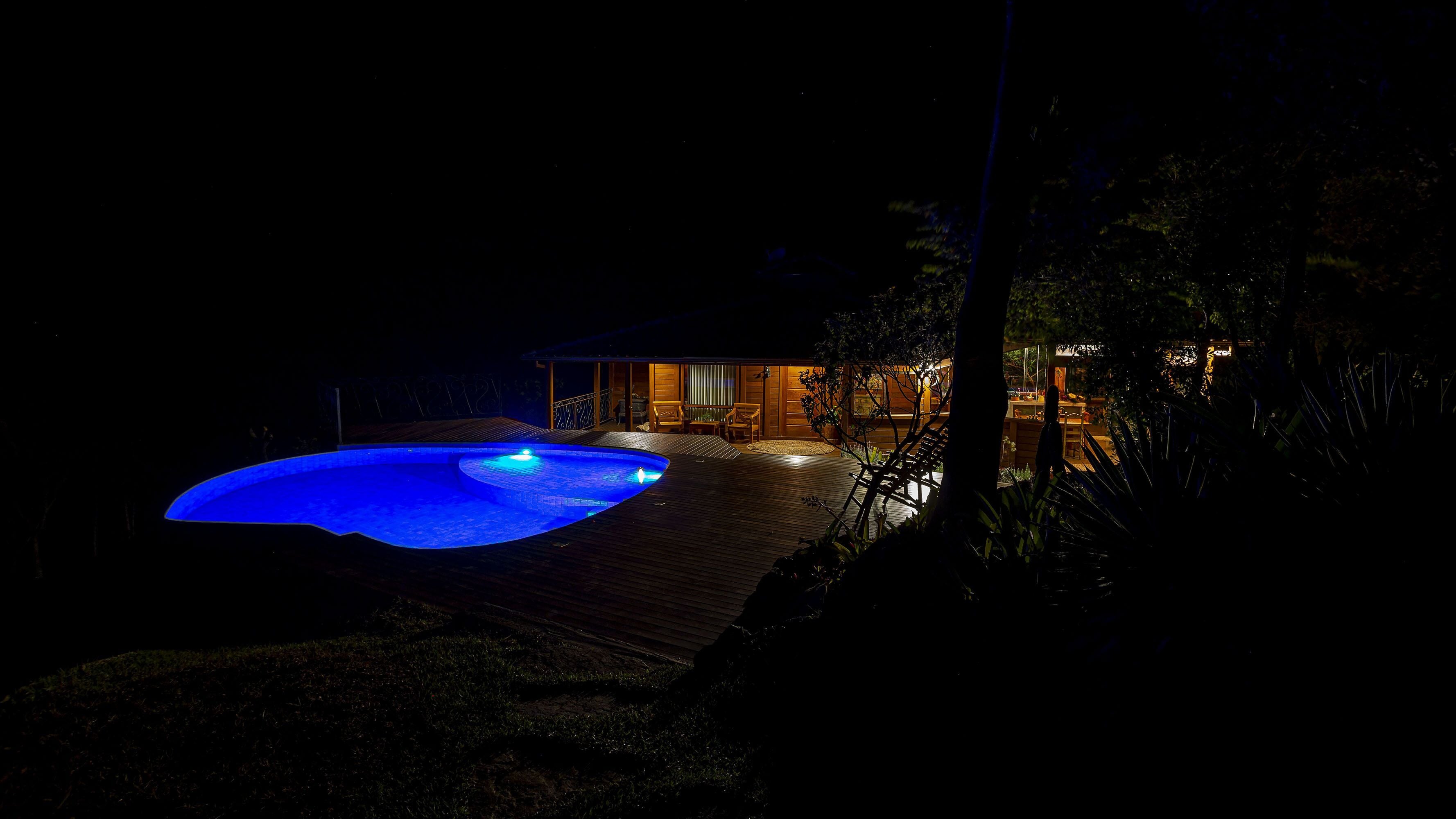 A nighttime view of a swimming pool with a wooden deck and a covered seating area.