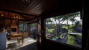 A cozy interior with a wooden ceiling, a brick fireplace, and a large window overlooking a landscaped garden. - Sitio Jatoba Lapinha da Serra (Santana do Riacho)