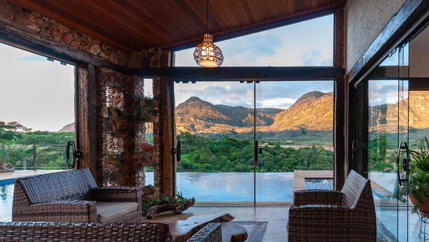 A modern room with wicker furniture, a stone wall, and a view of mountains and a lake. - Sitio Jatoba Lapinha da Serra (Santana do Riacho)