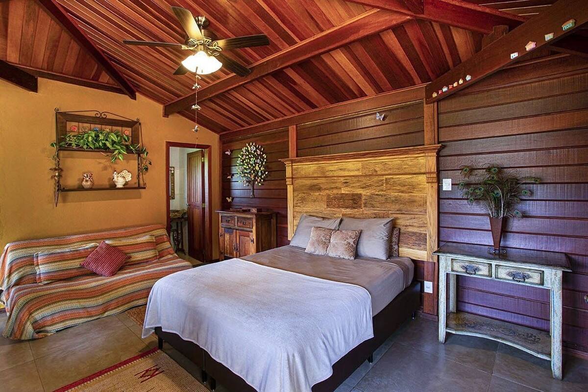 Une chambre avec un plafond en bois, un lit recouvert d’une courtepointe blanche, une table de chevet en bois et une étagère murale ornée de plantes.