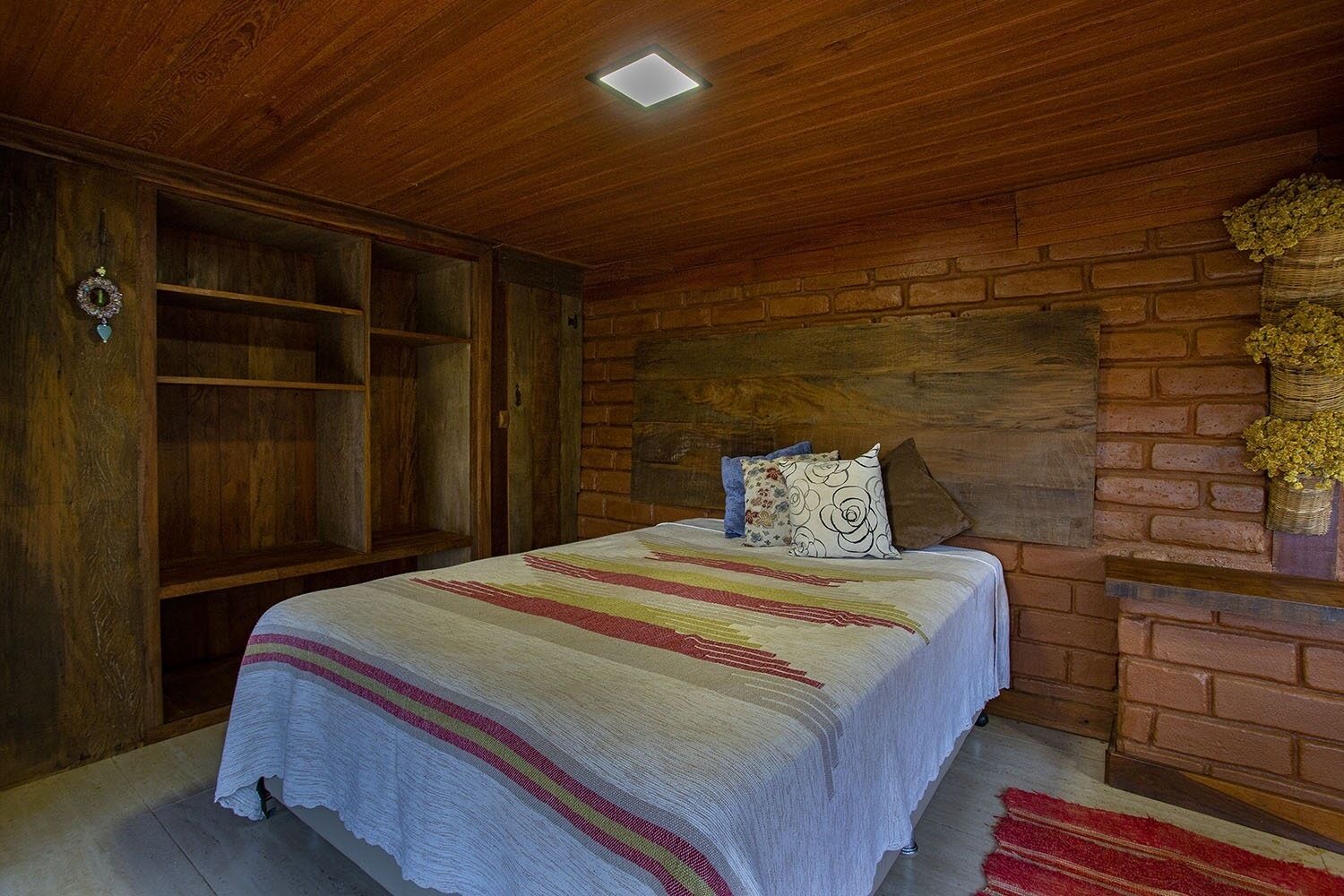 A bedroom with a wooden ceiling, a brick wall, a bed with a colorful blanket, and a small fireplace with flowers.