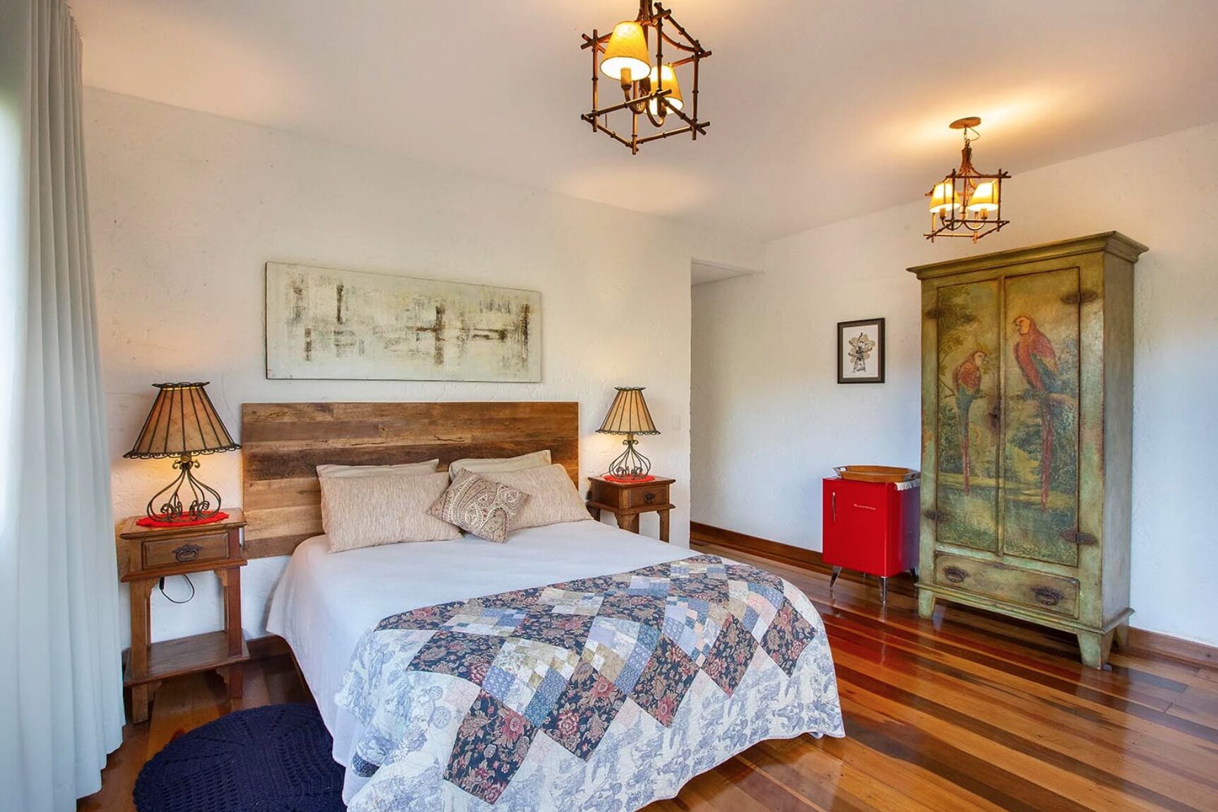 A bedroom with a wooden headboard, a quilted bedspread, two bedside lamps, a red cabinet, and a large painting on the wall.