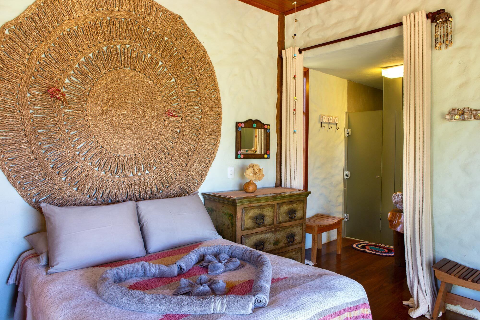 A bedroom with a large woven headboard, a wooden dresser, a mirror with colorful decorations, and a bed with a striped blanket.
