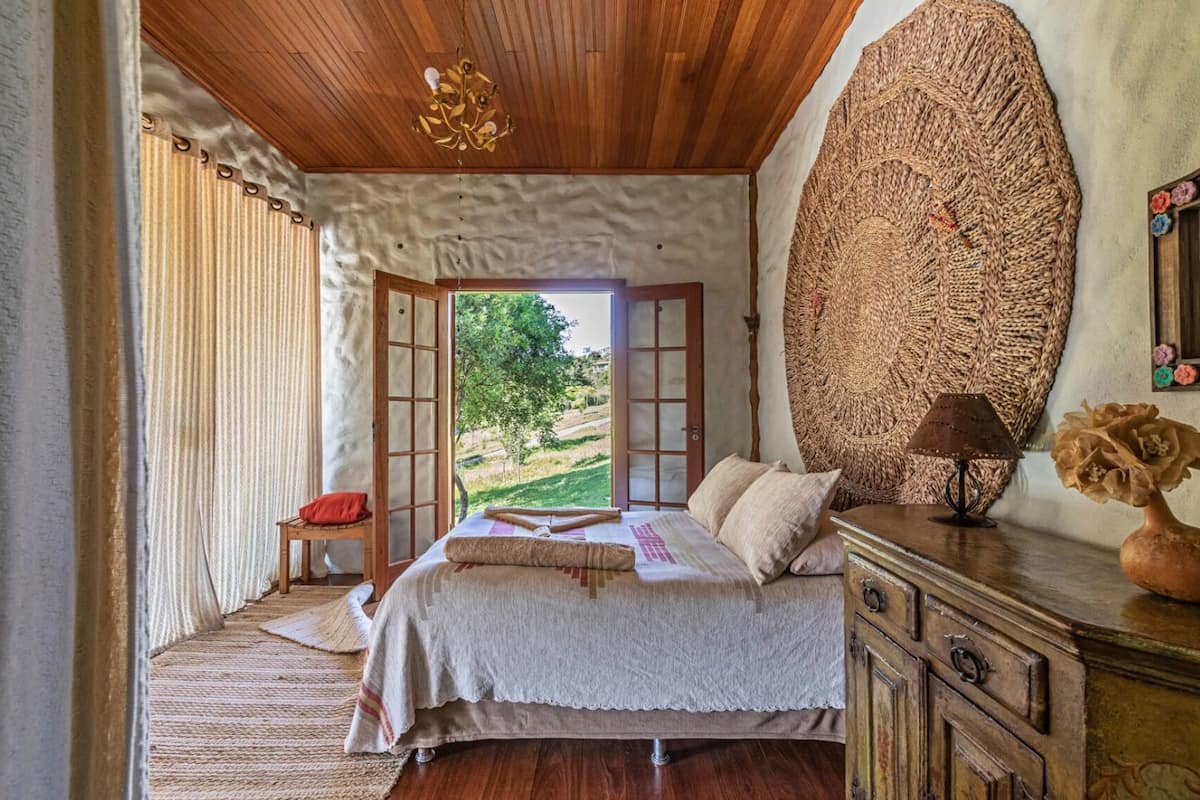 Une chambre avec un grand lit, une commode en bois, une lampe et une vue sur l’extérieur.