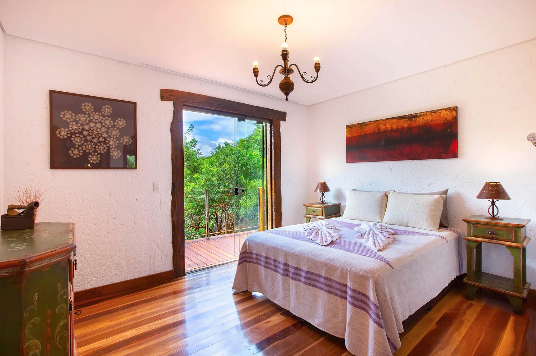 A bedroom with a large bed, a chandelier, a painting, and a view of greenery through a glass door.