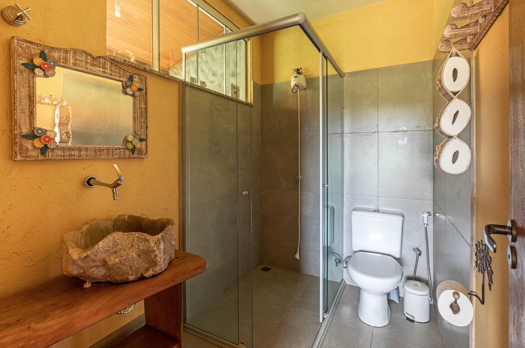 Un baño con lavamanos de piedra, cabina de ducha con paredes de vidrio, inodoro y espejo con detalles florales.