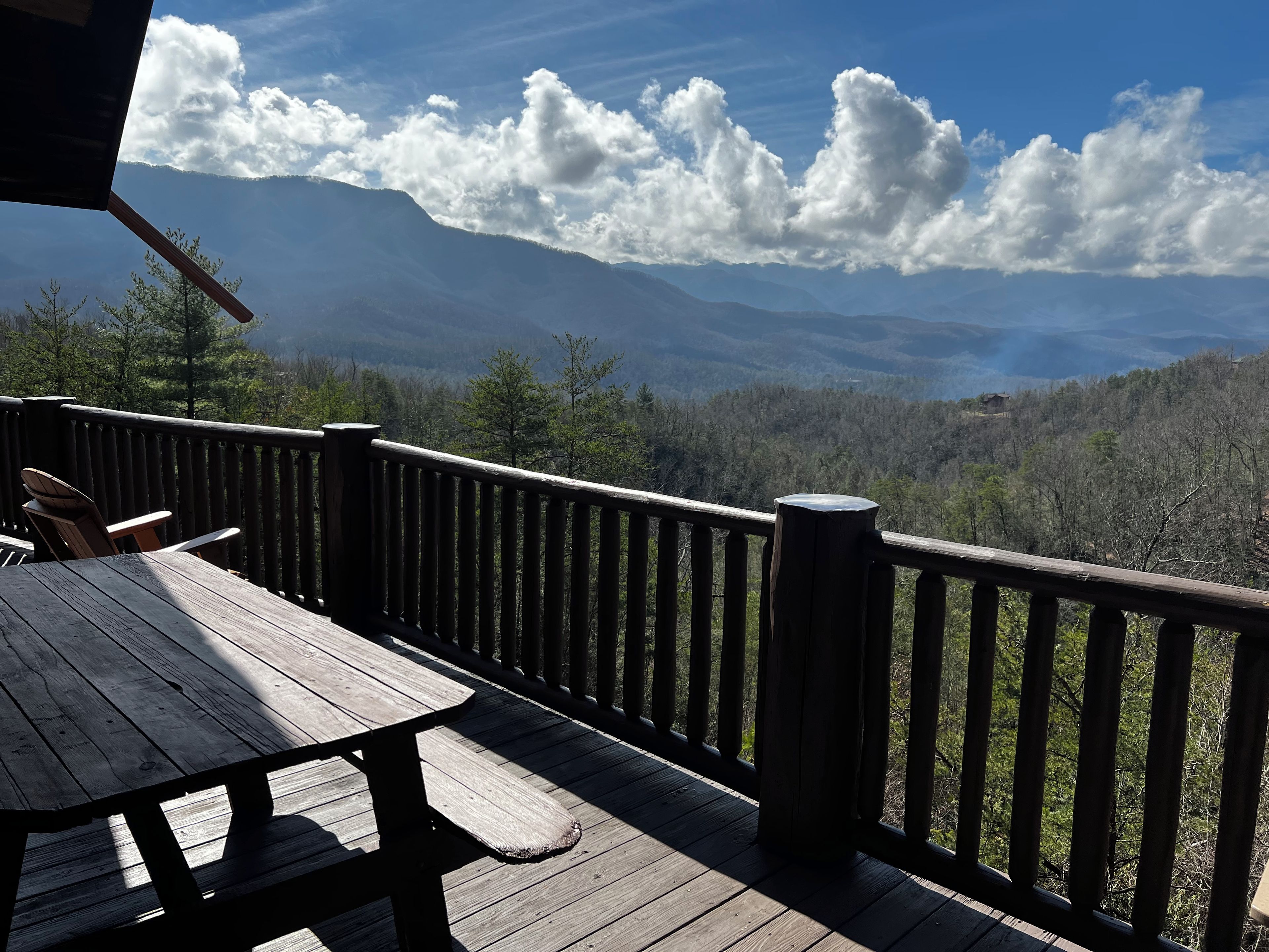Une terrasse en bois avec une table et des chaises, offrant une vue sur un paysage montagneux, avec des arbres et un ciel dégagé.