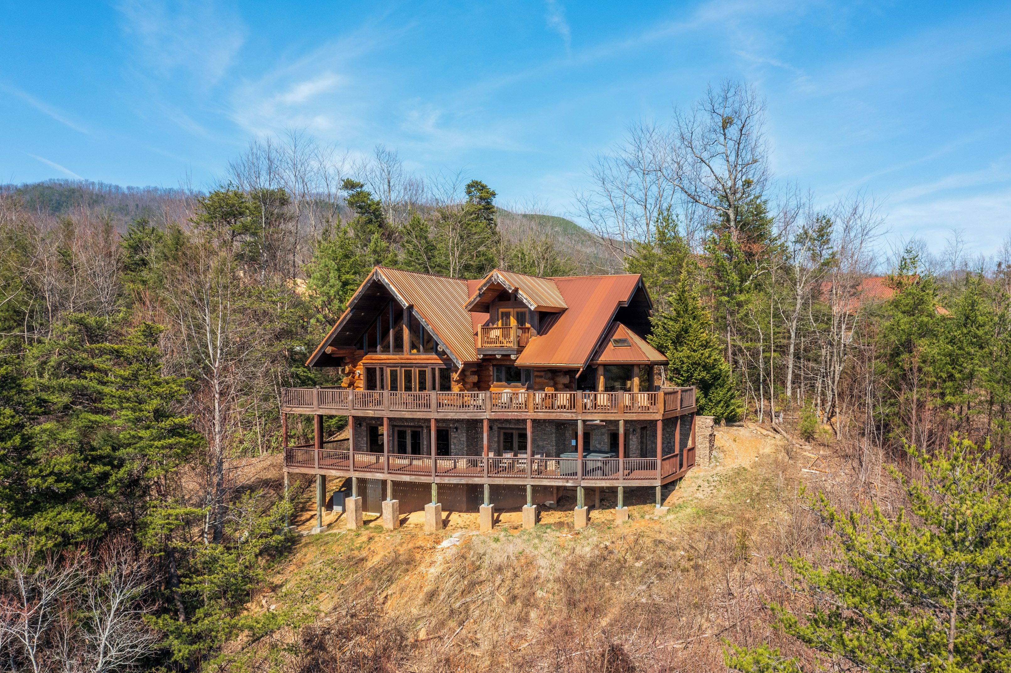 A large wooden cabin with a balcony, surrounded by trees and hills.