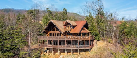 A large wooden cabin with a balcony, surrounded by trees and hills.