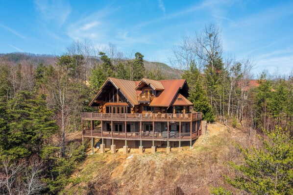 A large wooden cabin with a balcony, surrounded by trees and hills.