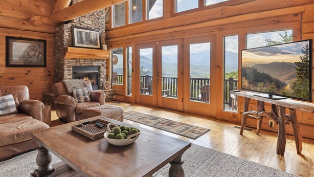 A cozy cabin interior with a stone fireplace, leather sofas, a wooden coffee table, and large windows offering mountain views.