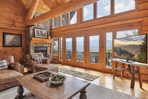 A cozy cabin interior with a stone fireplace, leather sofas, a wooden coffee table, and large windows offering mountain views. - Cabin w/Breathtaking Mtn. Views-Game Room-Hot Tub (Gatlinburg)