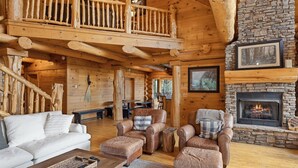 A log cabin interior with a fireplace, leather sofas, and a wooden staircase.