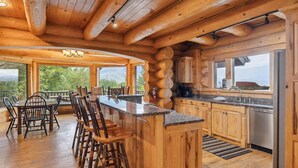 A log cabin interior with a bar area, wooden dining set, kitchen, and large windows offering scenic views.
