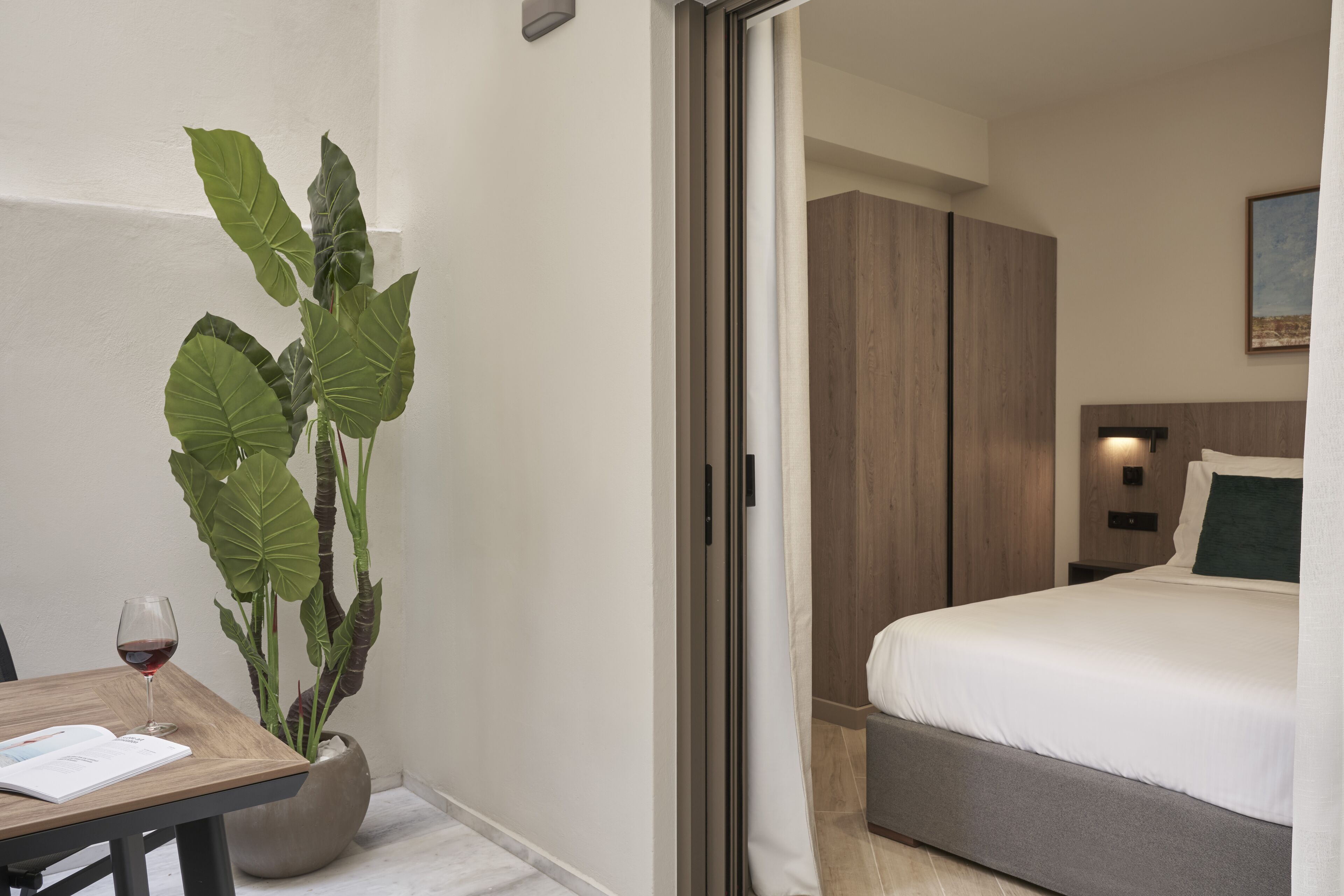 A hotel room with a bed, a bedside table with a glass of red wine, and a large potted plant.