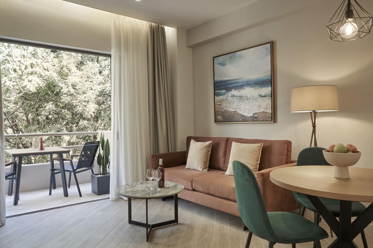 En moderne stue med sofa, sofabord og spiseplads. Der er en balkon med udsigt over træer og en strand.