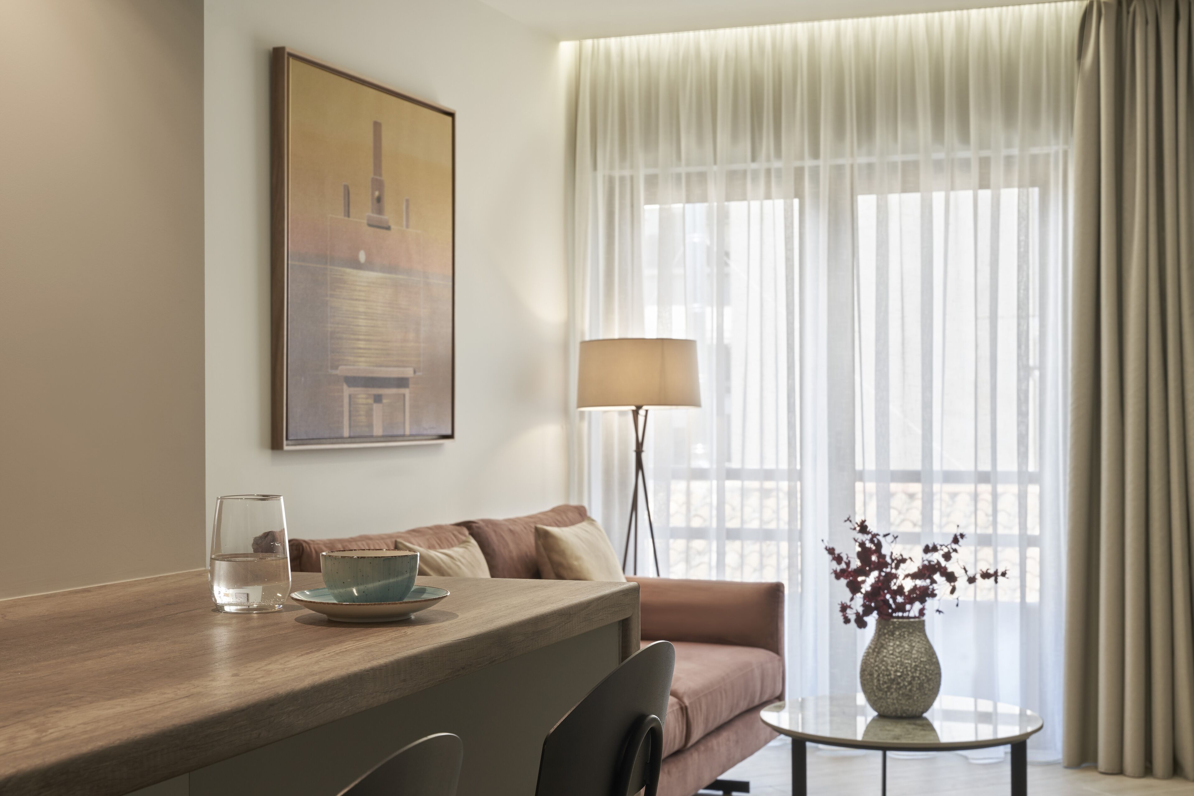 Een hotelkamer met een houten bureau, een bank, een vaas met bloemen en een schilderij aan de muur.