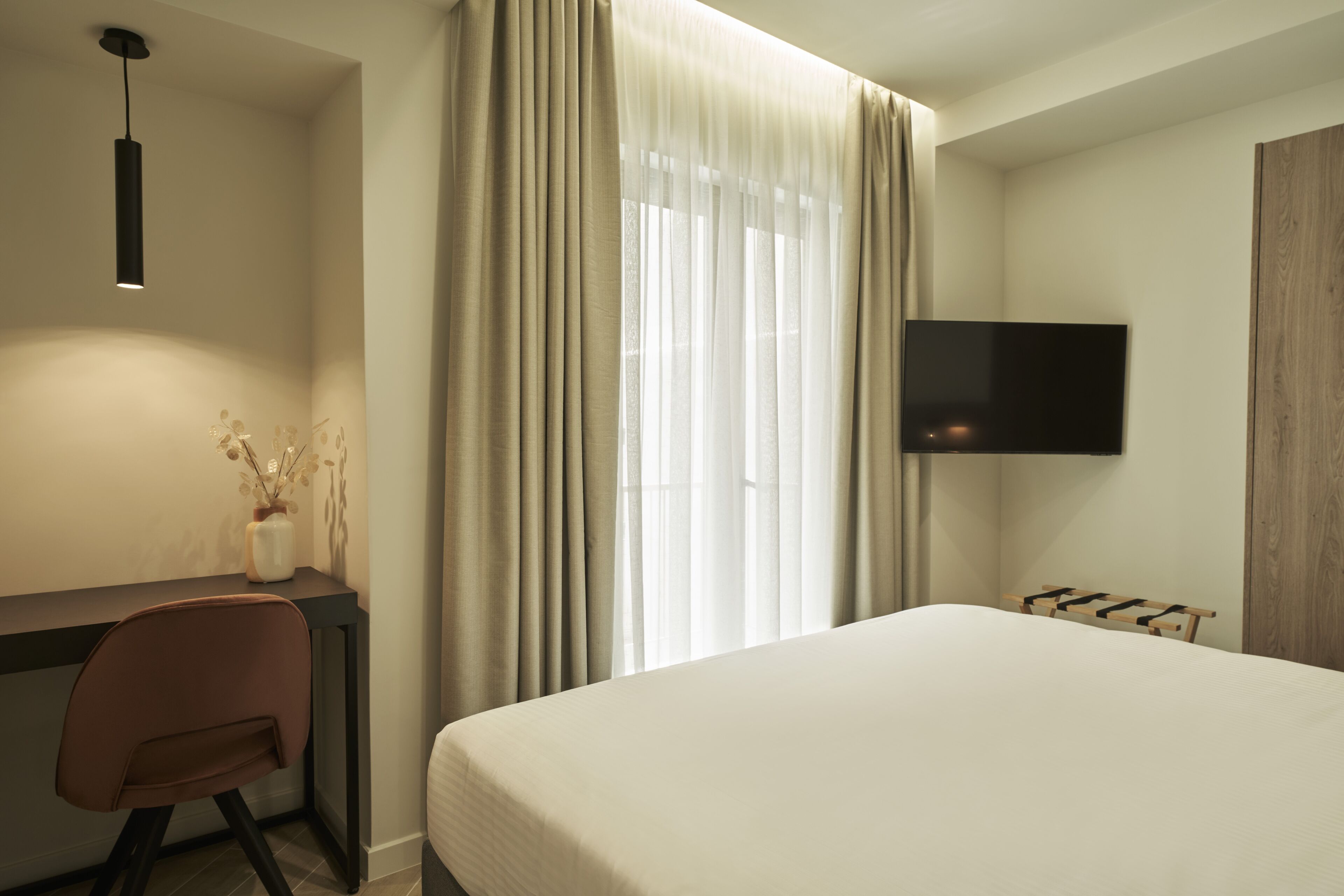 A hotel room with a bed, a desk with a chair, a TV mounted on the wall, and a window with curtains.