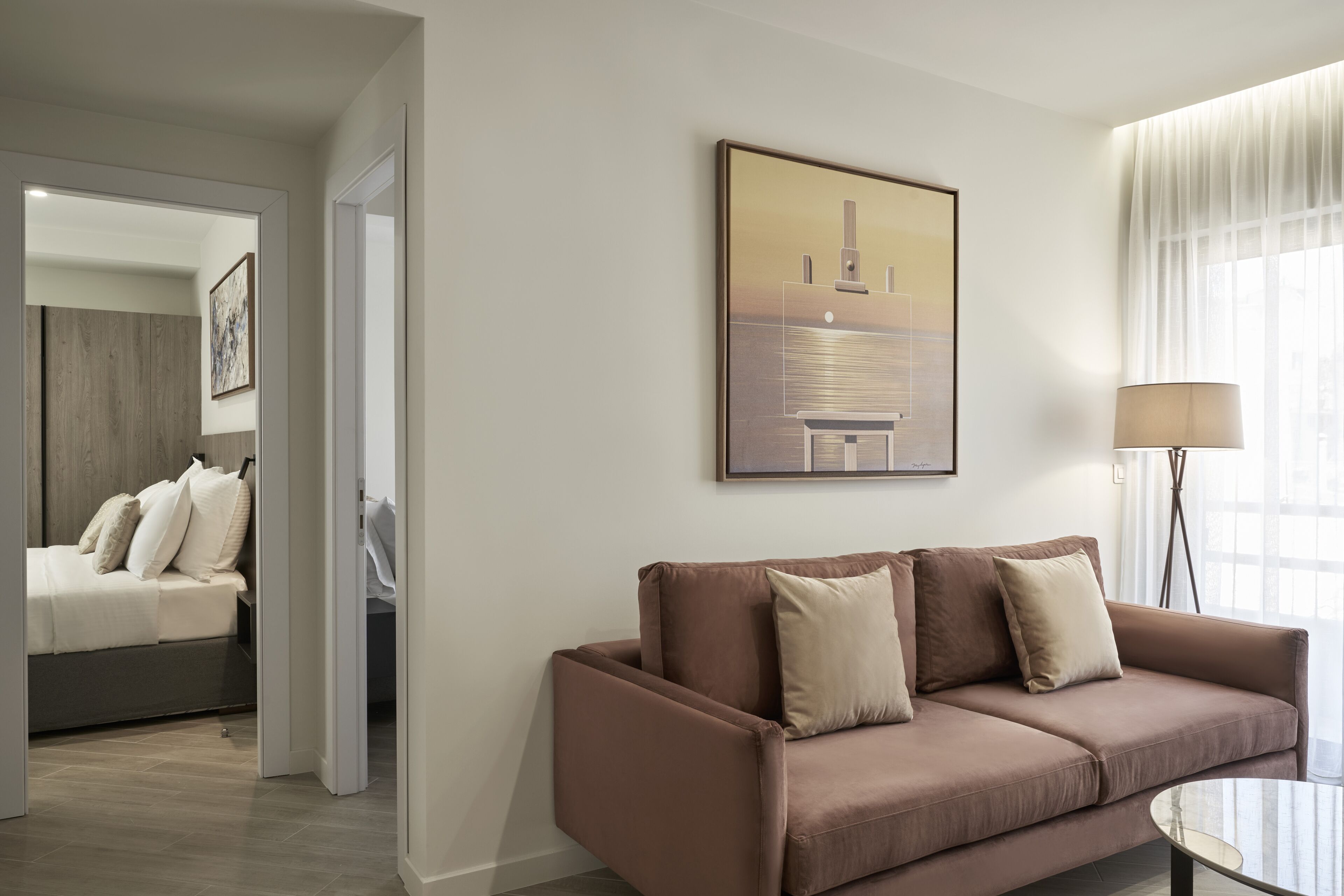 A modern hotel room with a brown sofa, a glass coffee table, a painting on the wall, and a bed with a wooden headboard.