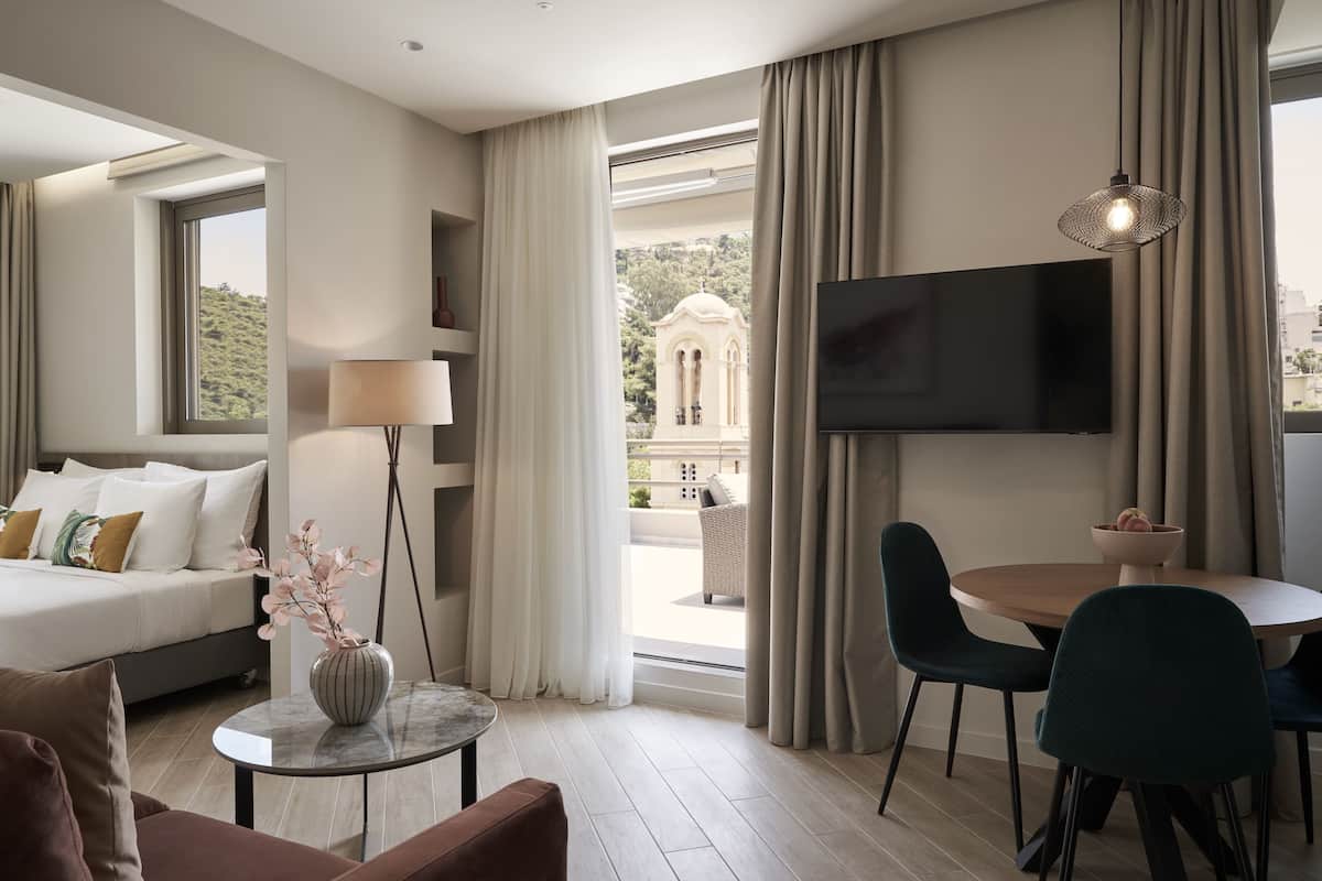 Et moderne hotelværelse med en sofa, et spisebord med stole, et fjernsyn og udsigt til en bygning gennem store vinduer.