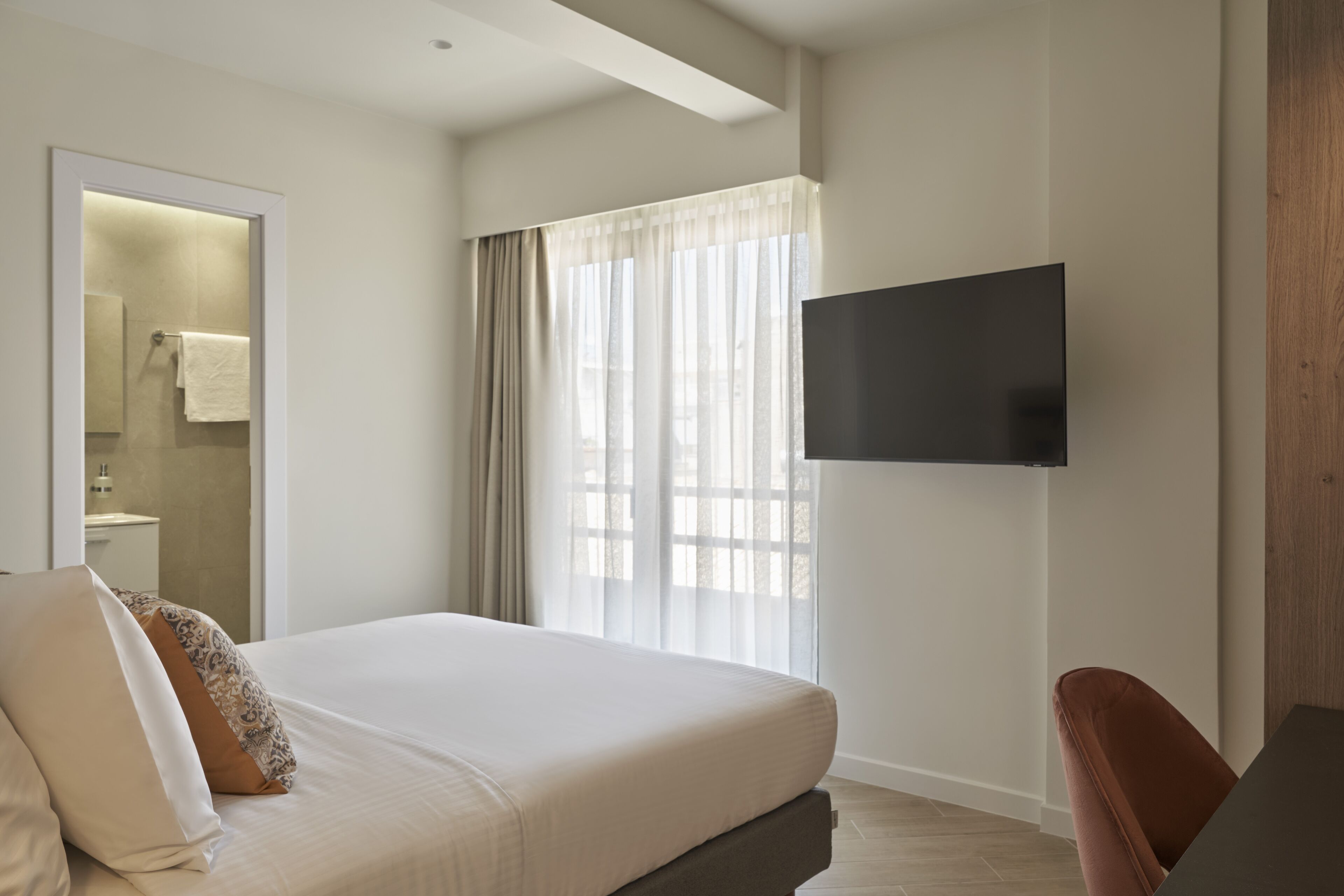 Ein modernes Hotelzimmer mit einem großen Bett, einem an der Wand befestigten Flachbildfernseher, einem Badezimmer mit Handtuchhalter und einem Fenster mit durchsichtigen Vorhängen.
