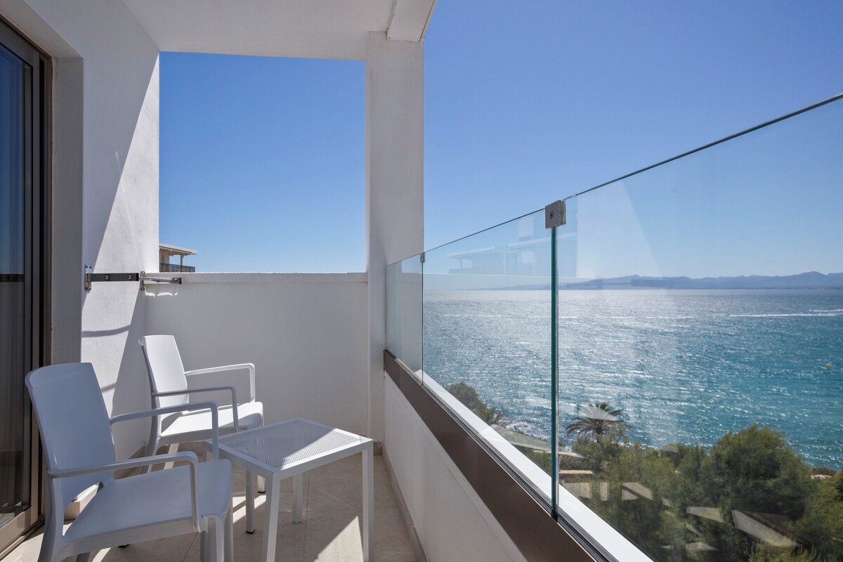 Un balcón con sillas y una mesa blancos, con vistas a un cuerpo de agua y tierra a lo lejos.