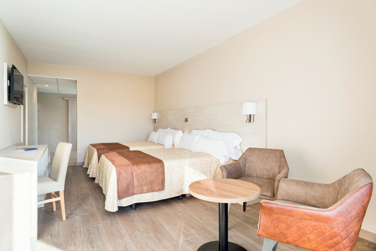 A hotel room with a bed, two armchairs, a small table, a desk, and a TV mounted on the wall.