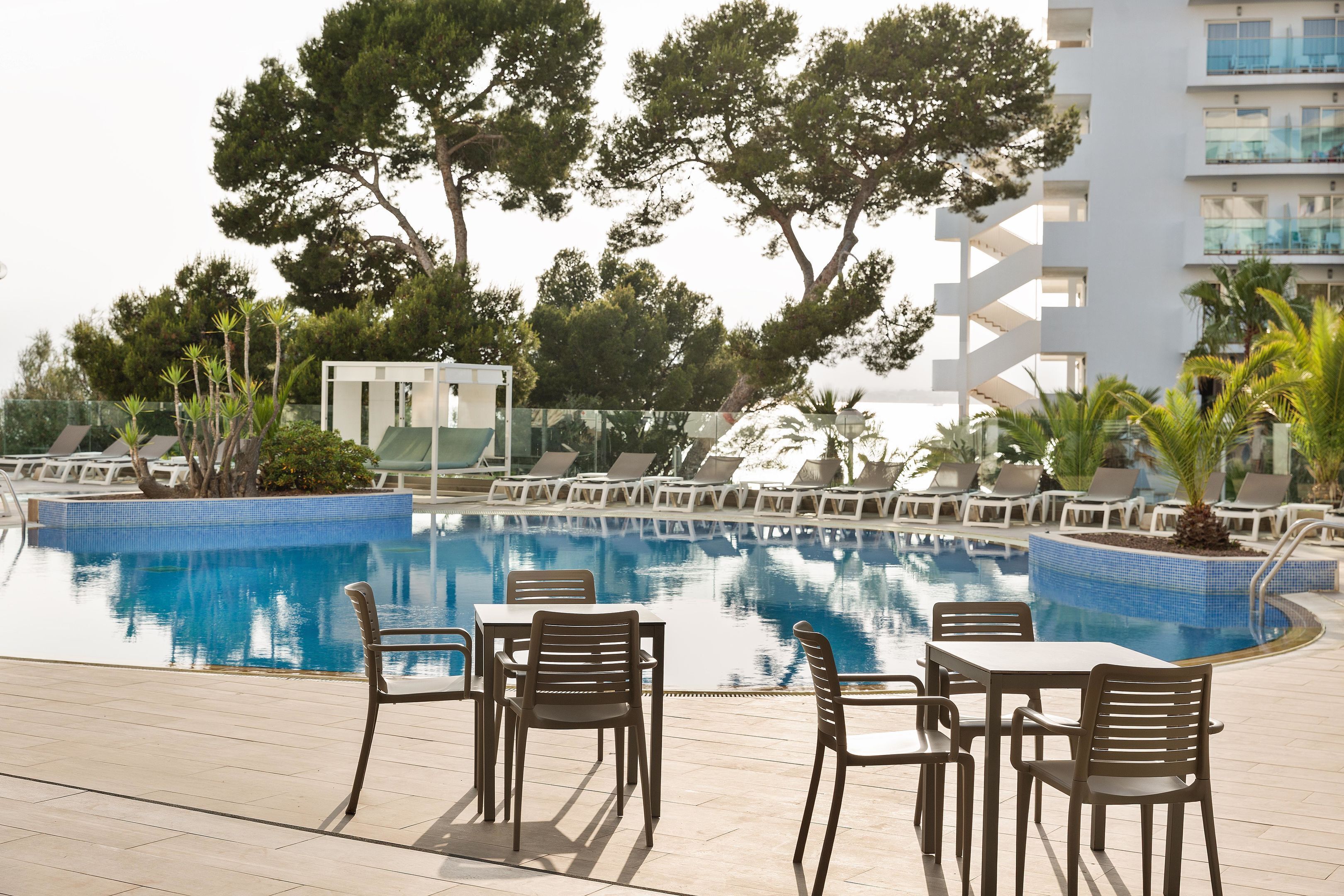 A pool area with lounge chairs and tables, surrounded by trees and a modern building.