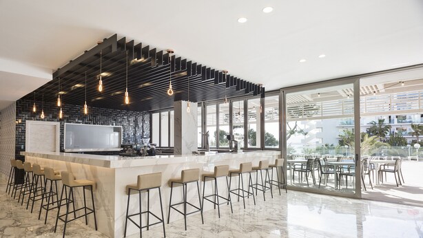 A modern bar area with a marble countertop, bar stools, pendant lighting, and large windows offering views of the outdoors. - Hotel Best Punta Dorada (Salou)