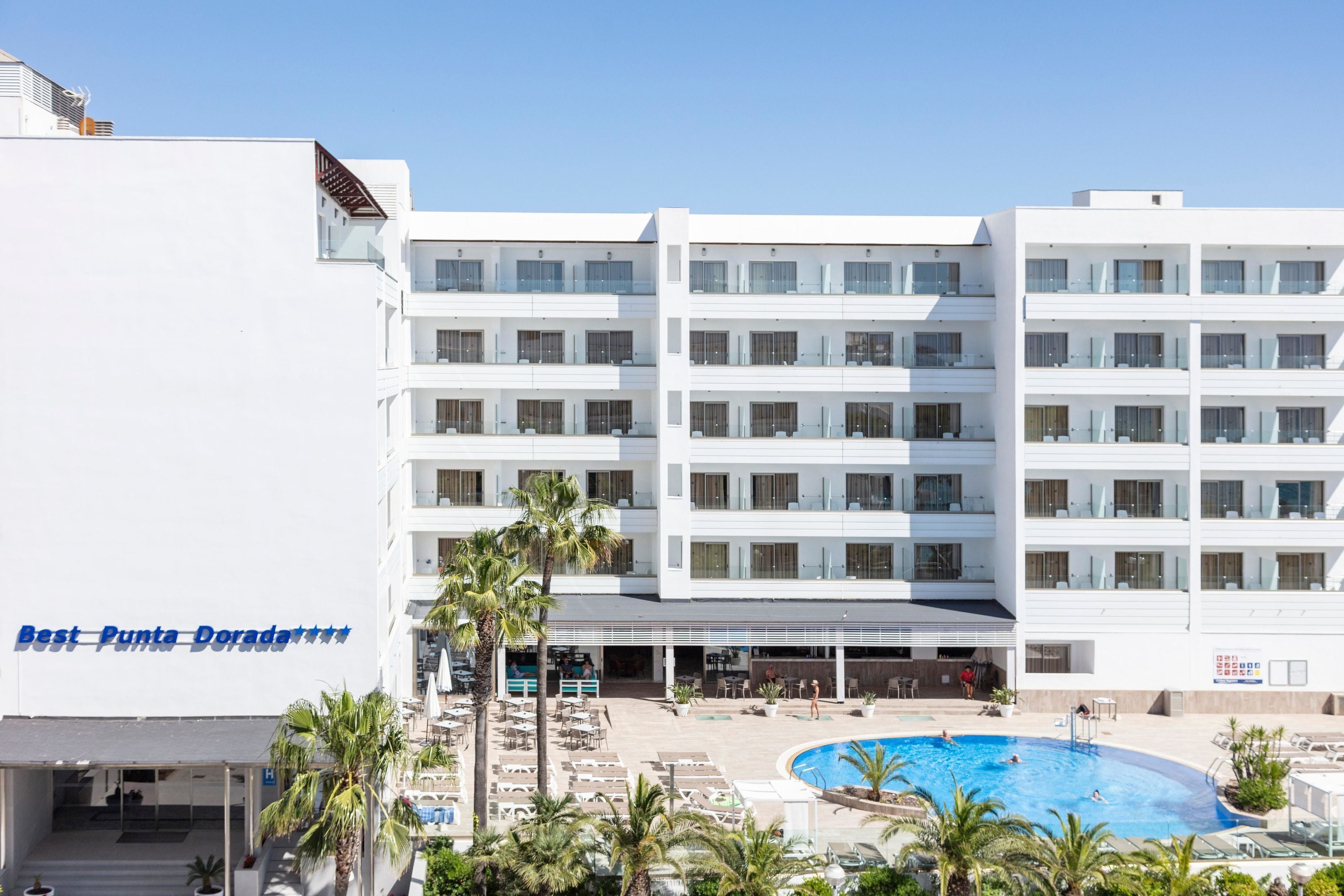 Een meerlaags hotel met een zwembad, palmbomen en een helderblauwe lucht.