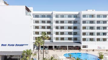 Een meerlaags hotel met een zwembad, palmbomen en een helderblauwe lucht.
