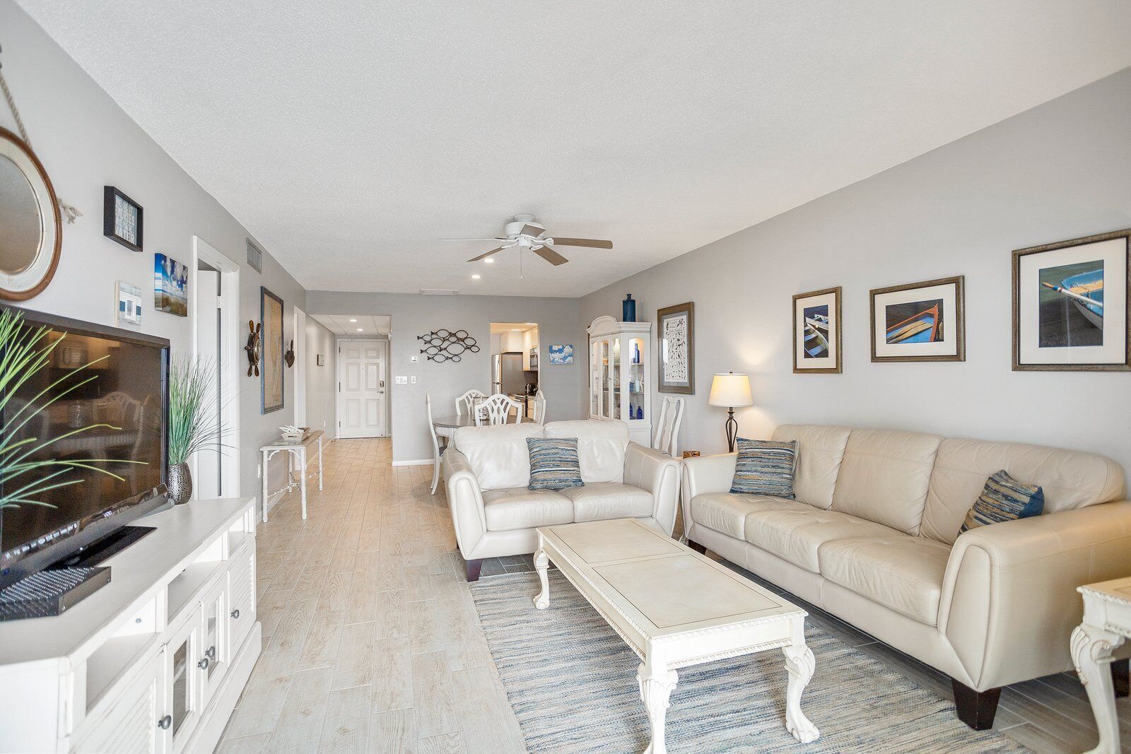 A spacious living room with a beige sofa, a white coffee table, and a TV on a white cabinet. There is a ceiling fan, a dining area with a white table and chairs, and a kitchen in the background.