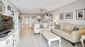 Un vaste salon avec un canapé beige, une table basse blanche et un téléviseur posé sur un meuble blanc. On y trouve également un ventilateur de plafond, un coin repas avec une table et des chaises blanches, et une cuisine à l’arrière-plan.
