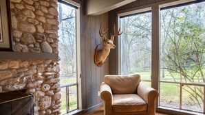 Una habitación con pared de piedra, cabeza de venado montada, un sillón beige y ventanas amplias que dan a un paisaje de árboles.