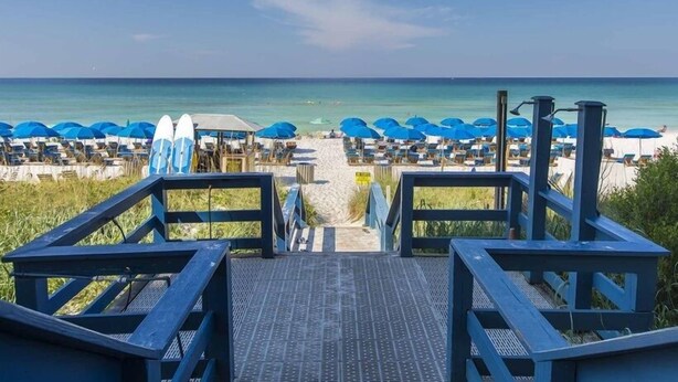 Blick auf einen Strand mit einer Holzpromenade, die zum Strand führt, Reihen blauer Sonnenschirme und Surfbrettern.