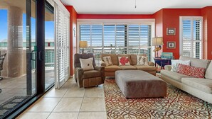 Una sala de estar con sofá, sillón y reposapiés, ventanas amplias con persianas y vistas al exterior.