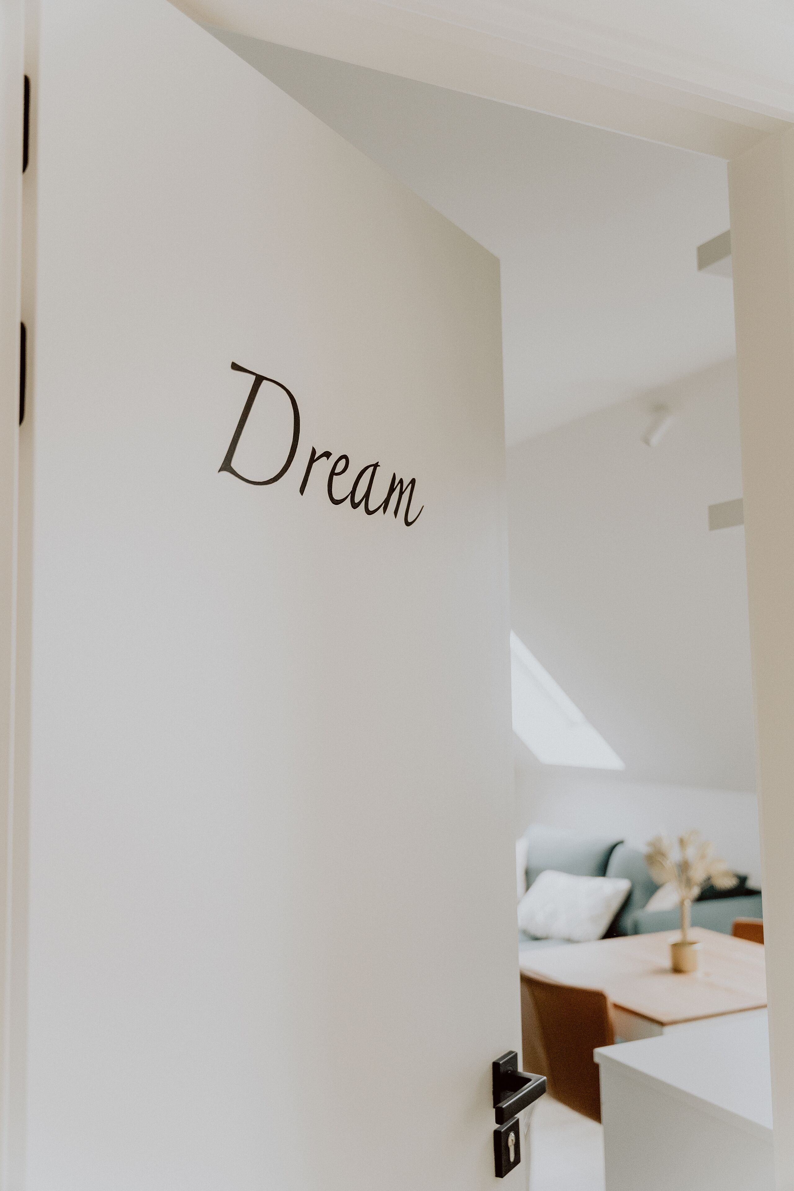 A door with the word "Dream" on it, leading into a room with a bed, a table, and a plant.