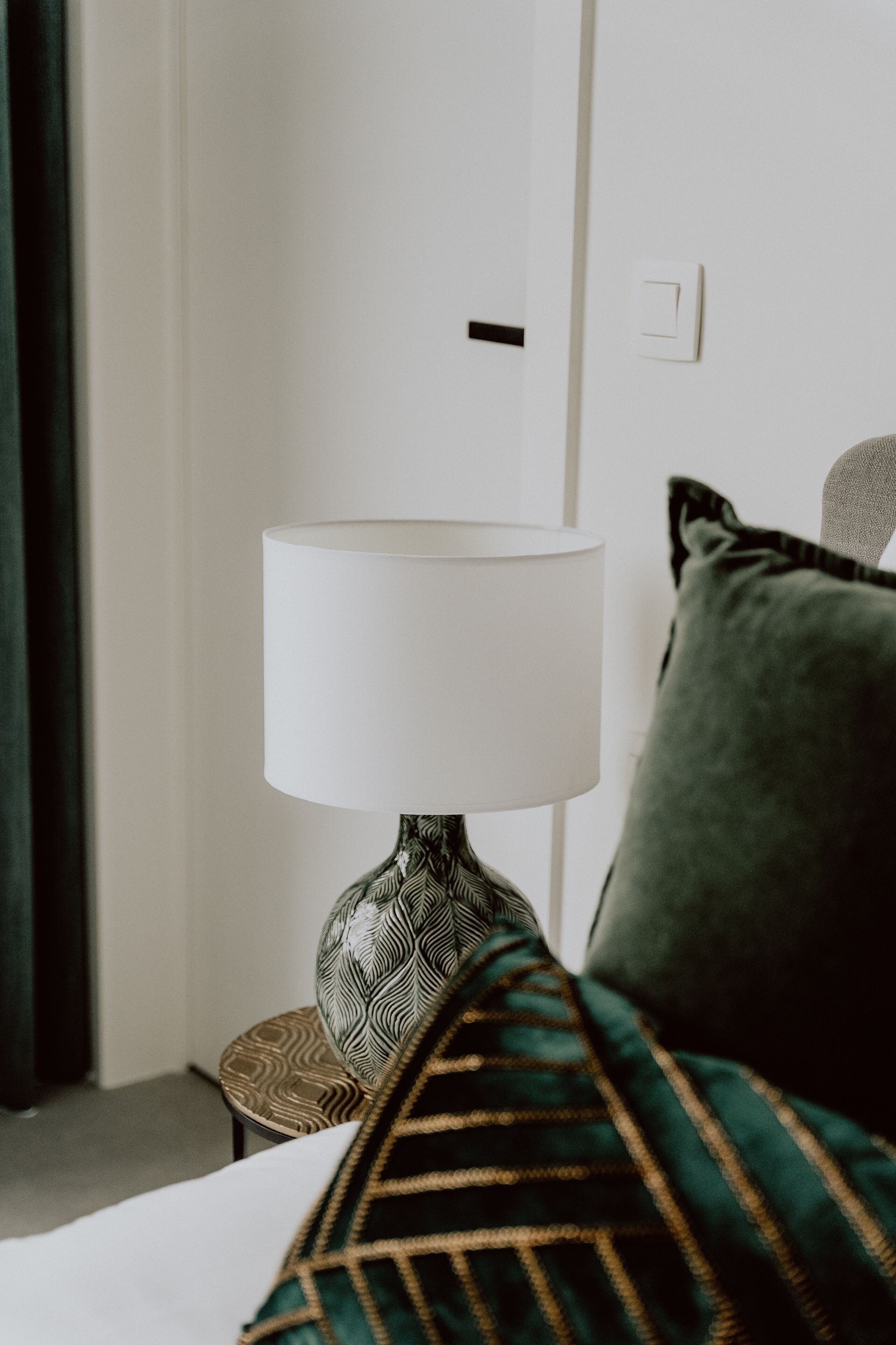 Een netjes opgemaakt bed met een deken in groen en goud, een witte lamp met een decoratieve voet en een donkergroen kussen op een kleine ronde stoel.