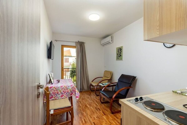 Habitació petita amb terra de fusta, una taula de dinar amb caires, una cuina reduïda amb fogó i una finestra amb cortines.