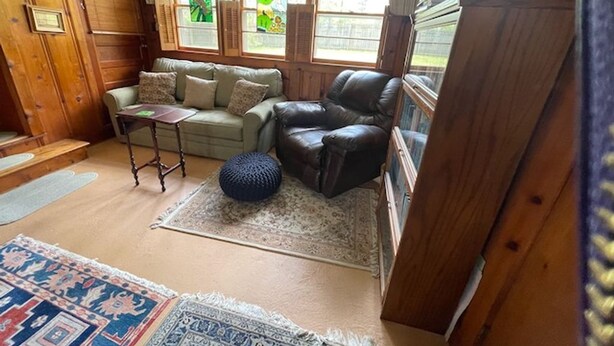 Een gezellige woonkamer met een bank, fauteuil en voetenbankje, houten wanden en grote ramen met gordijnen.