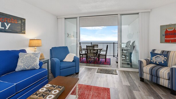 A living room with a blue sofa, a striped armchair, and a coffee table. There is a glass door leading to a balcony with a view of the ocean. - Pyramid 11J by Seaside Vacations & Sales (Ocean City)