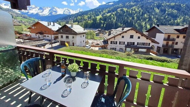 Un balcon avec une table dressée pour deux personnes, donnant sur un village de montagne aux bâtiments traditionnels et offrant une vue sur des sommets enneigés.