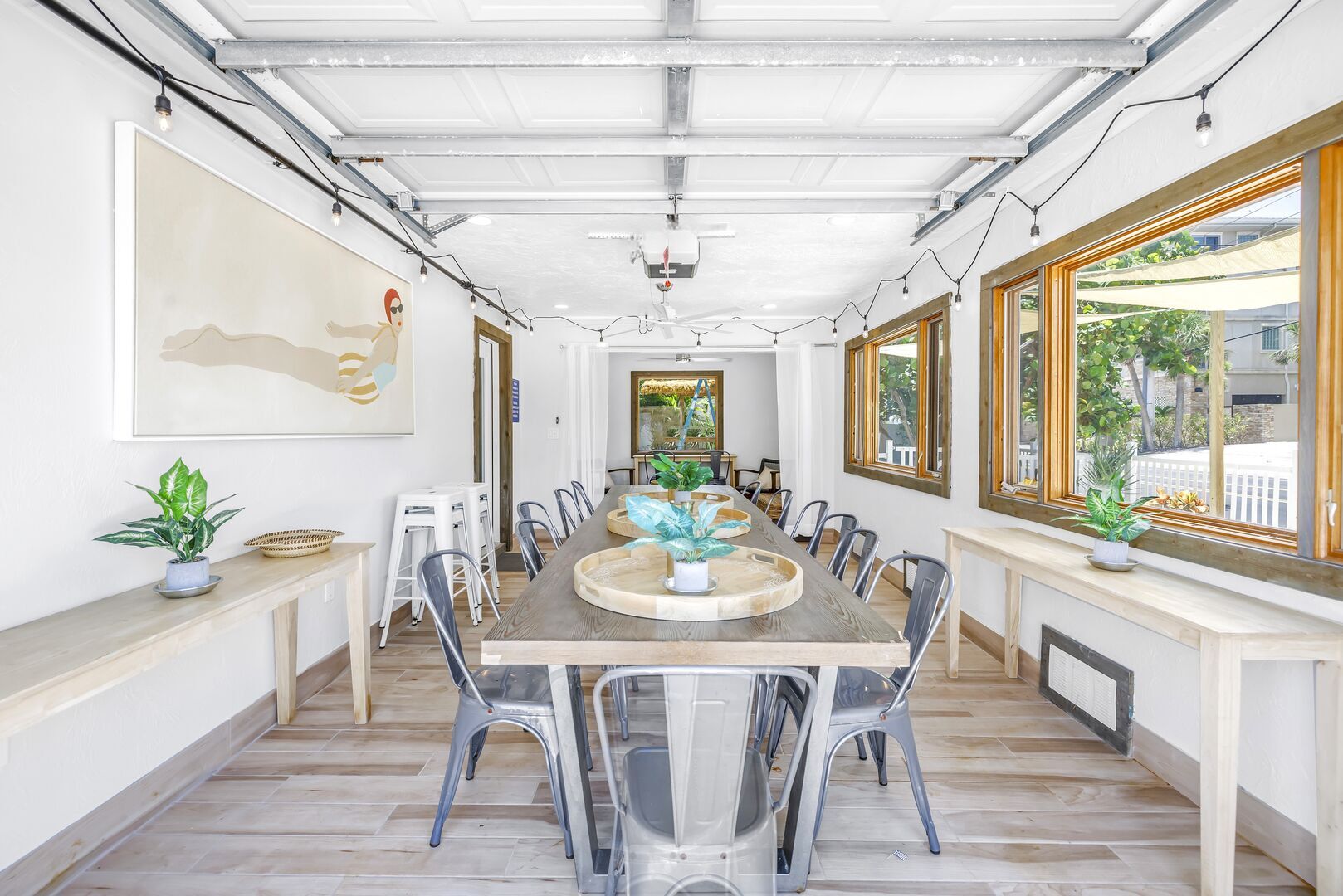 A spacious dining area with a long wooden table, clear plastic chairs, and potted plants.