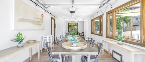 A spacious dining area with a long wooden table, clear plastic chairs, and potted plants.