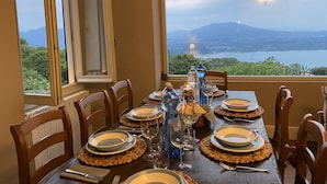 A dining table set for six with plates, glasses, and a candle. The table is positioned near a window with a view of a lake and mountains.