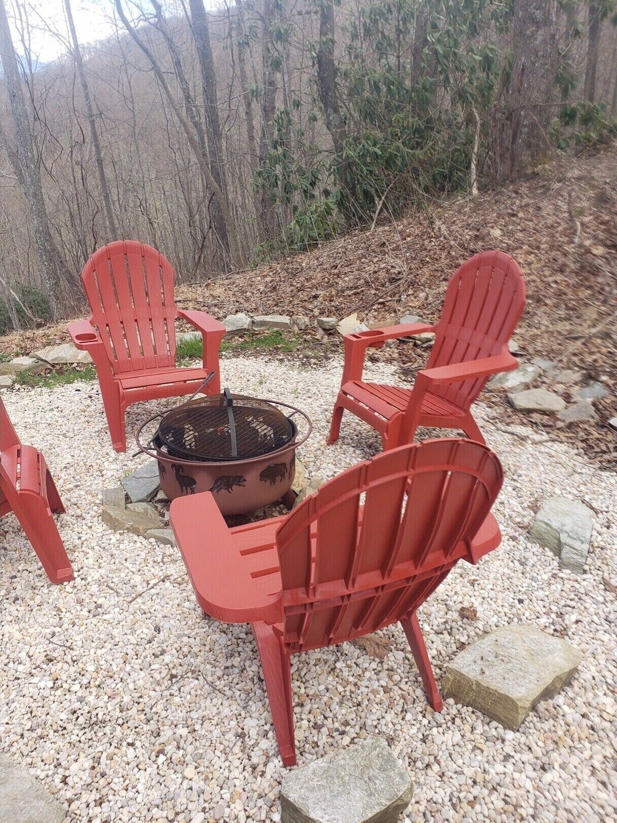 A group of four red Adirondack chairs arranged around a fire pit in a forested area.