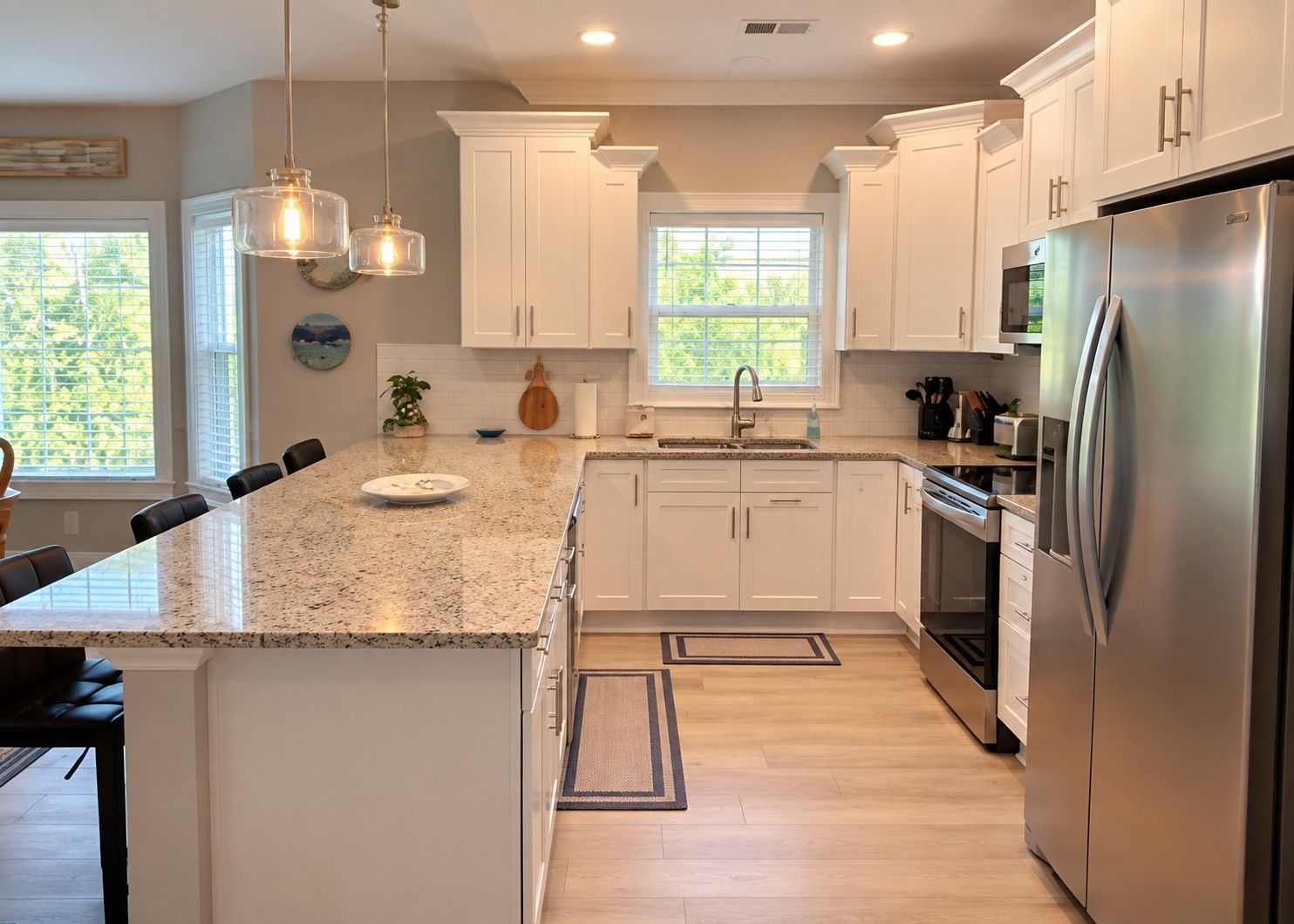 A modern kitchen with white cabinets, a granite countertop, stainless steel appliances, pendant lighting, and a view of greenery outside.